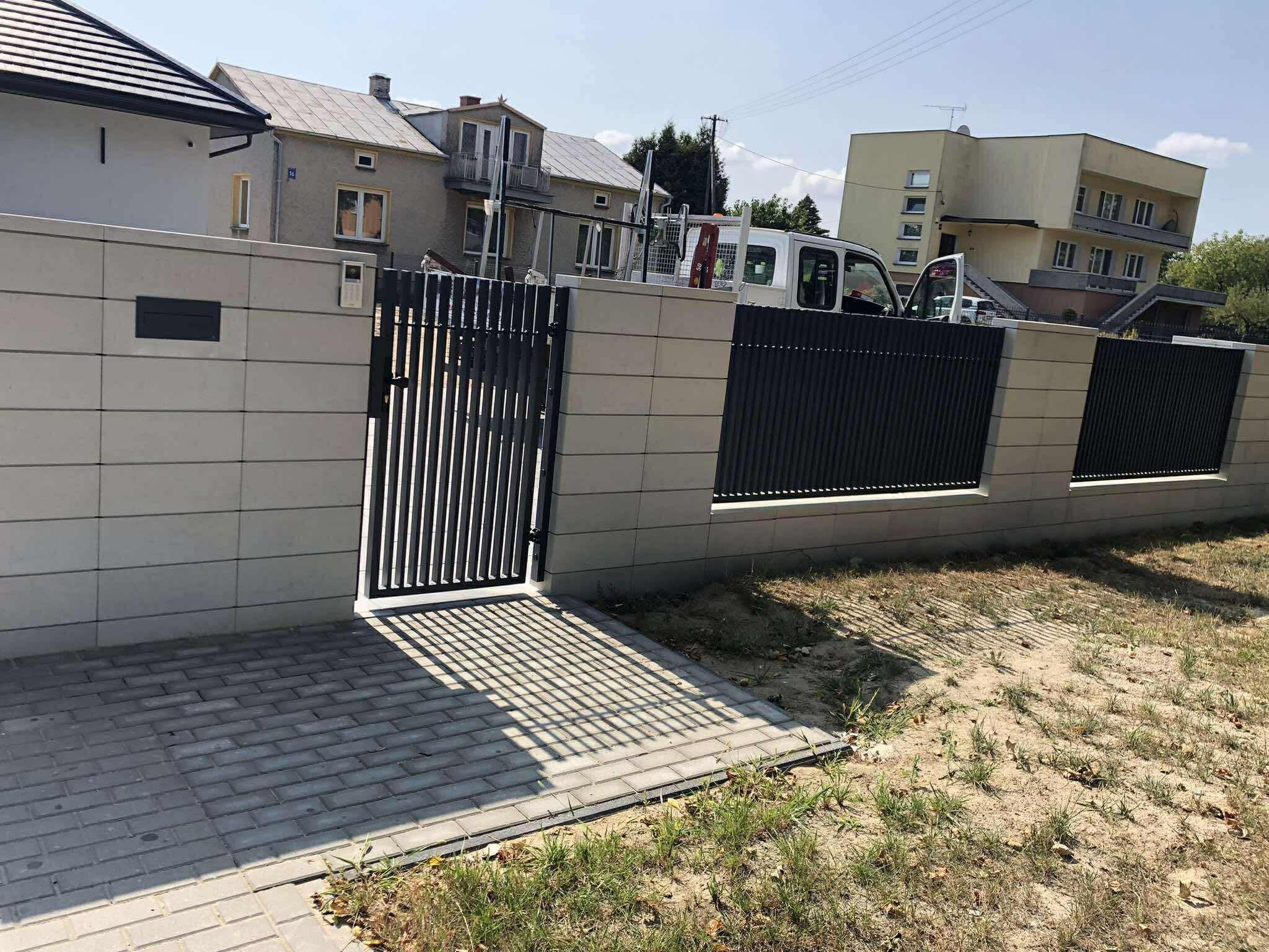 Nowoczesne ogrodzenie panelowe z bramką furtką, z szarej kostki betonowej, z widocznym domem w tle. Furtka rzuca cień na kostkę. Widok na ogrodzenie z zewnątrz posesji.