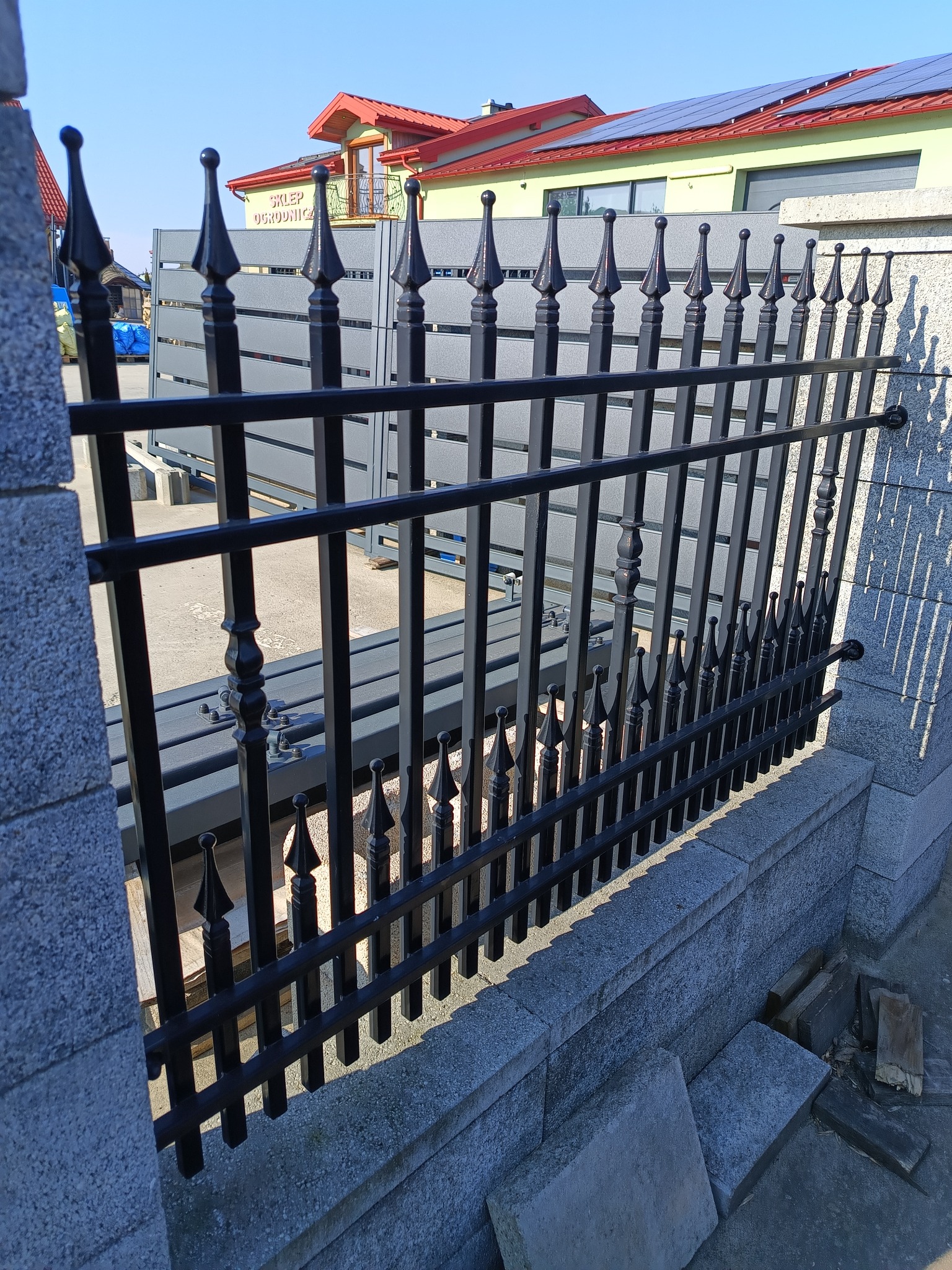 Czarne, metalowe ogrodzenie kute z ozdobnymi grotami, zamontowane na betonowej podmurówce. W tle widoczny budynek z czerwonym dachem i panelami słonecznymi.