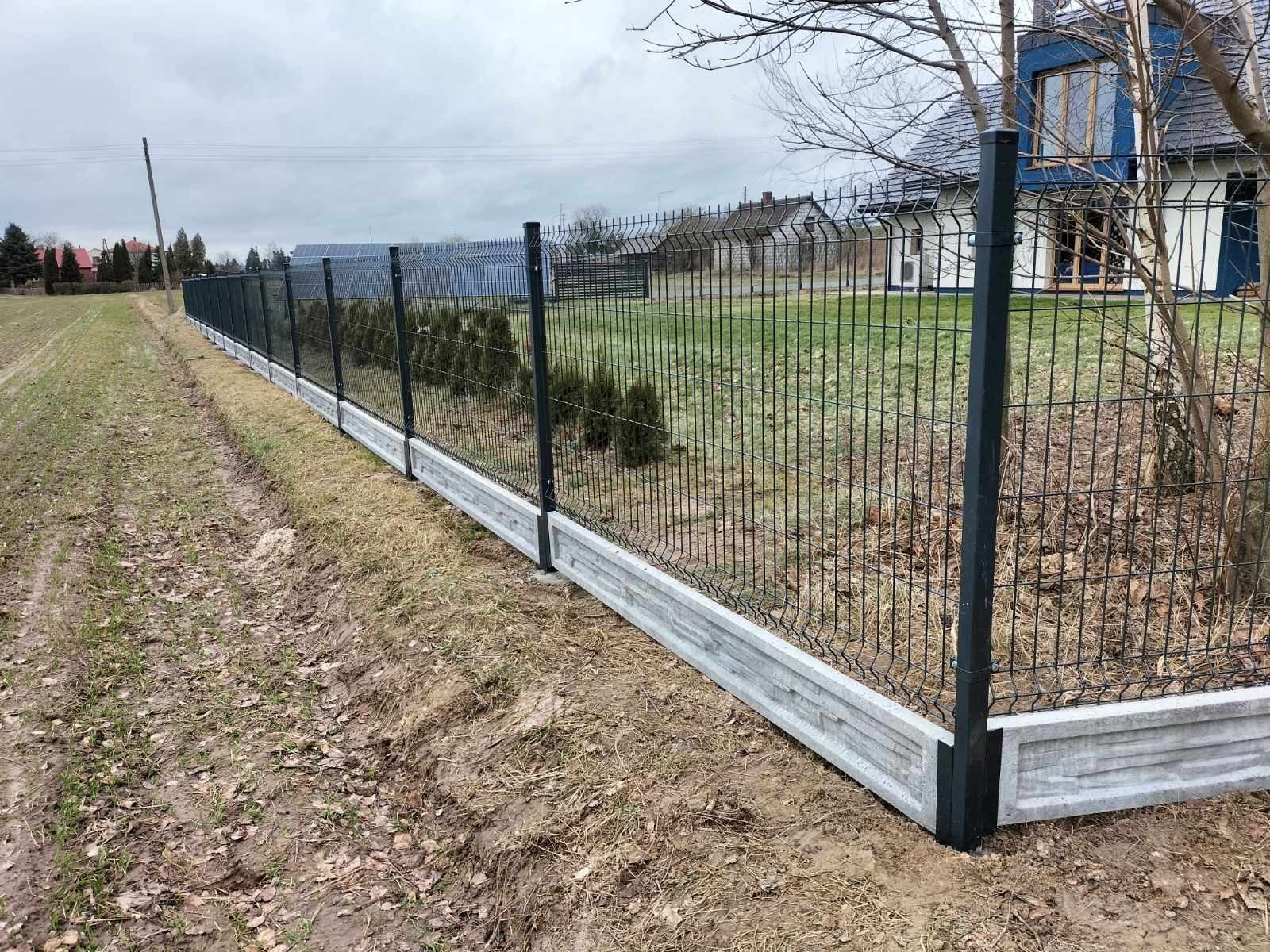 Panelowe ogrodzenie z podmurówką betonową na posesji z zielenią. Ciemne słupki i siatka kontrastują z jasną podmurówką. Widok z perspektywy ziemi.