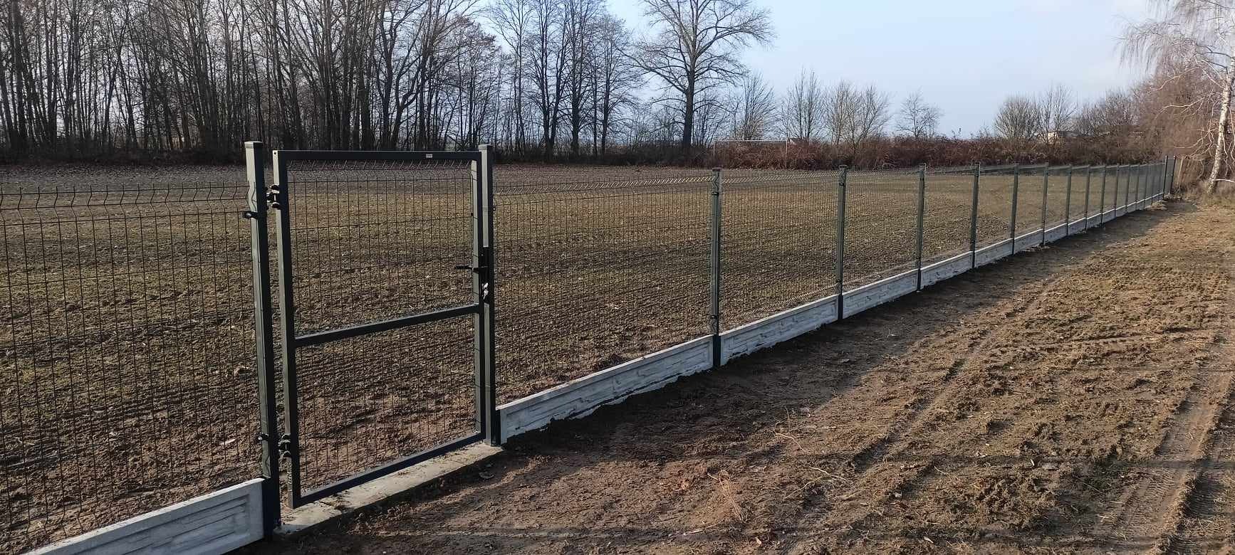 Panelowe ogrodzenie z bramą wjazdową, osadzone na betonowej podmurówce. Widoczne metalowe słupki i siatka ogrodzeniowa. Tło stanowią drzewa i pole.
