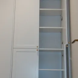 Wysoka, biała szafa z uchylnymi drzwiami, z otwartą częścią ukazującą puste półki, złote okucia, widok z bliska.