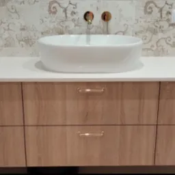 Nowoczesna łazienka z drewnianą szafką pod umywalką nablatową, wykończoną złotymi uchwytami i armaturą, na tle dekoracyjnych płytek.