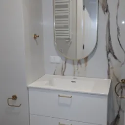 Biała szafka łazienkowa z dwiema szufladami, umywalką nablatową i owalnym lustrem z złotą ramą, oświetlona kulistymi lampami na ścianie z marmurowym wzorem płytek.