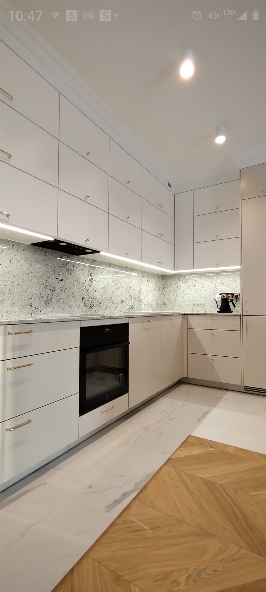 Nowoczesna kuchnia z białymi, gładkimi frontami szafek, złotymi uchwytami, blatem z jasnego kamienia z ciemnymi żyłkami i oświetleniem LED podszafkowym, widoczny piekarnik i drewniana podłoga...