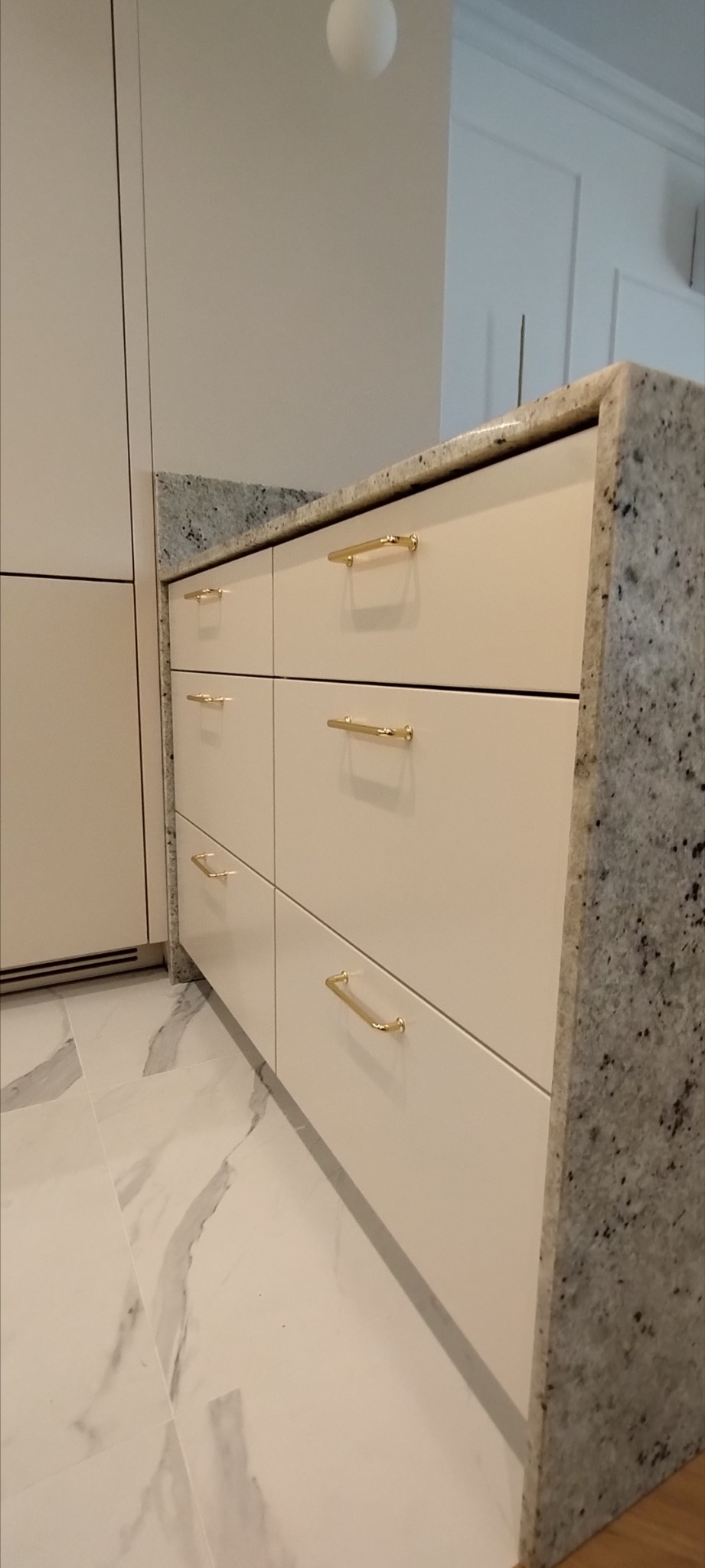 Nowoczesne, jasne szafki kuchenne z białymi frontami i złotymi uchwytami, wykończone blatem z szarego granitu o wyraźnej fakturze, na tle marmurowej podłogi.