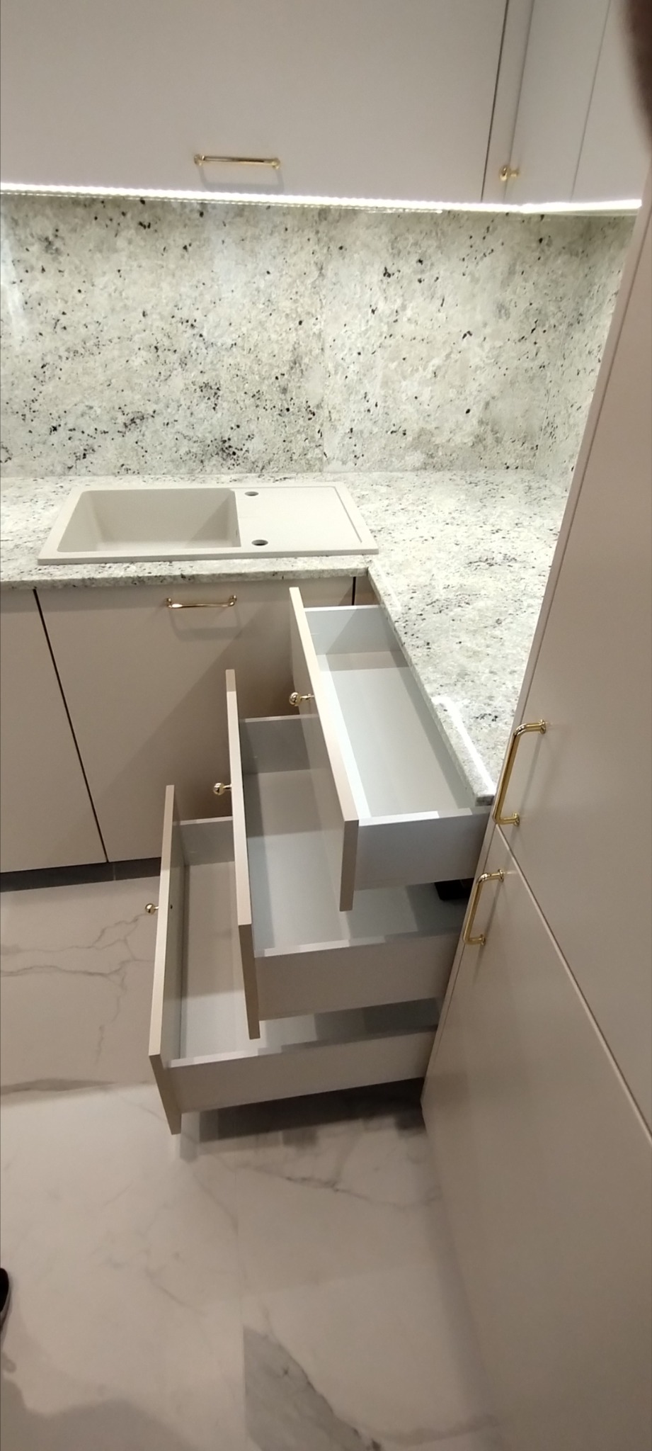 Jasna kuchnia na wymiar z otwartymi szufladami i blatem z jasnego kamienia z ciemnymi plamkami, widoczny zlew wbudowany w blat.