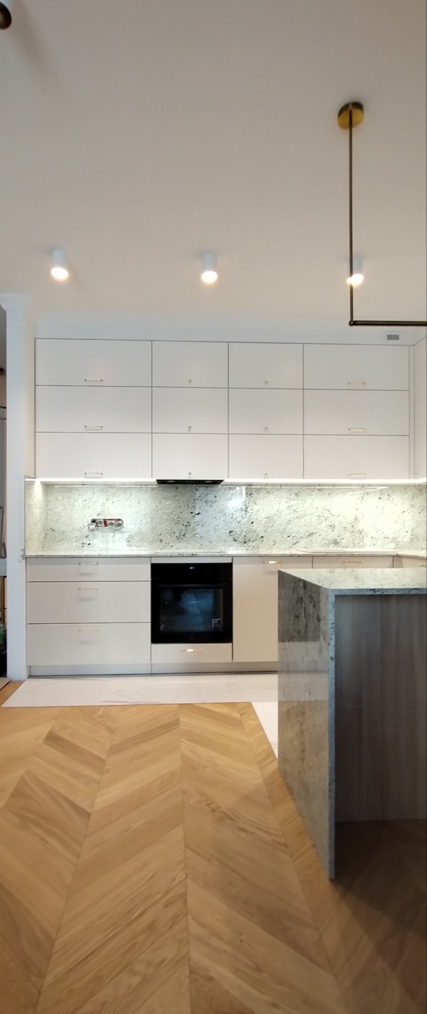 Nowoczesna, jasna kuchnia z białymi szafkami, blatem z jasnego kamienia i parkietem ułożonym w jodełkę, oświetlona punktowymi lampami i złotą wiszącą lampą.