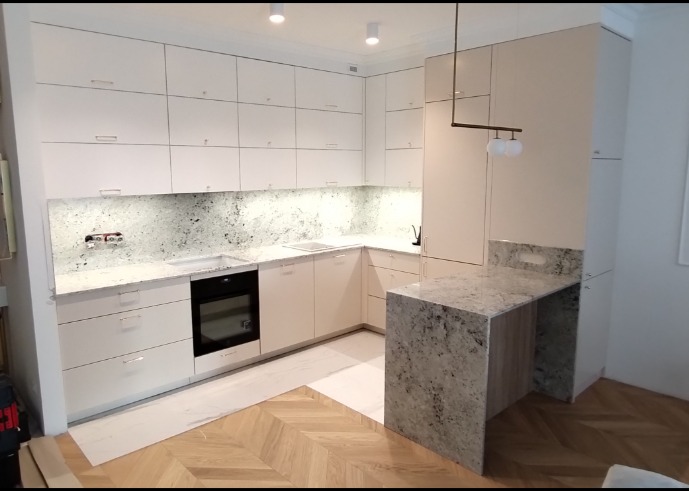 Jasna kuchnia z białymi szafkami, blatem z szarego granitu i drewnianą podłogą ułożoną w jodełkę; oświetlenie sufitowe i lampa wisząca nad barkiem.