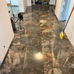 Marcin Florczuk - Połyskująca, brązowo-szara podłoga z płytek imitujących marmur, świeżo po ułożeniu, z widocznym odkurzaczem, drabiną, psem rasy Shiba Inu i żółtą zmiotką w tle remontowanego mieszkania.
