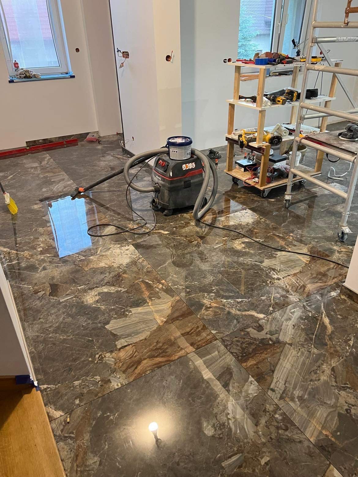 Polerowane, marmurowe płytki podłogowe w trakcie montażu, widoczny odkurzacz przemysłowy i narzędzia na wózku.