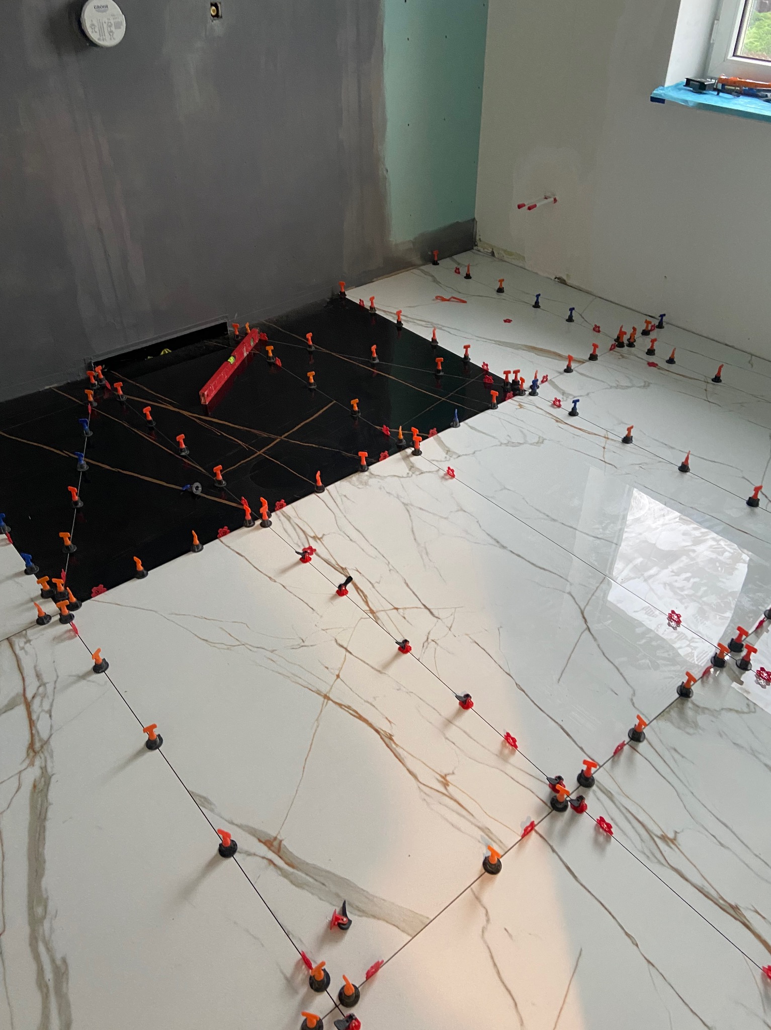 Częściowo ułożona podłoga z płytek, z czarnymi i białymi płytkami imitującymi marmur, użyto klipsów poziomujących i sznurków do wyrównania, widoczny poziomica na czarnych płytkach.