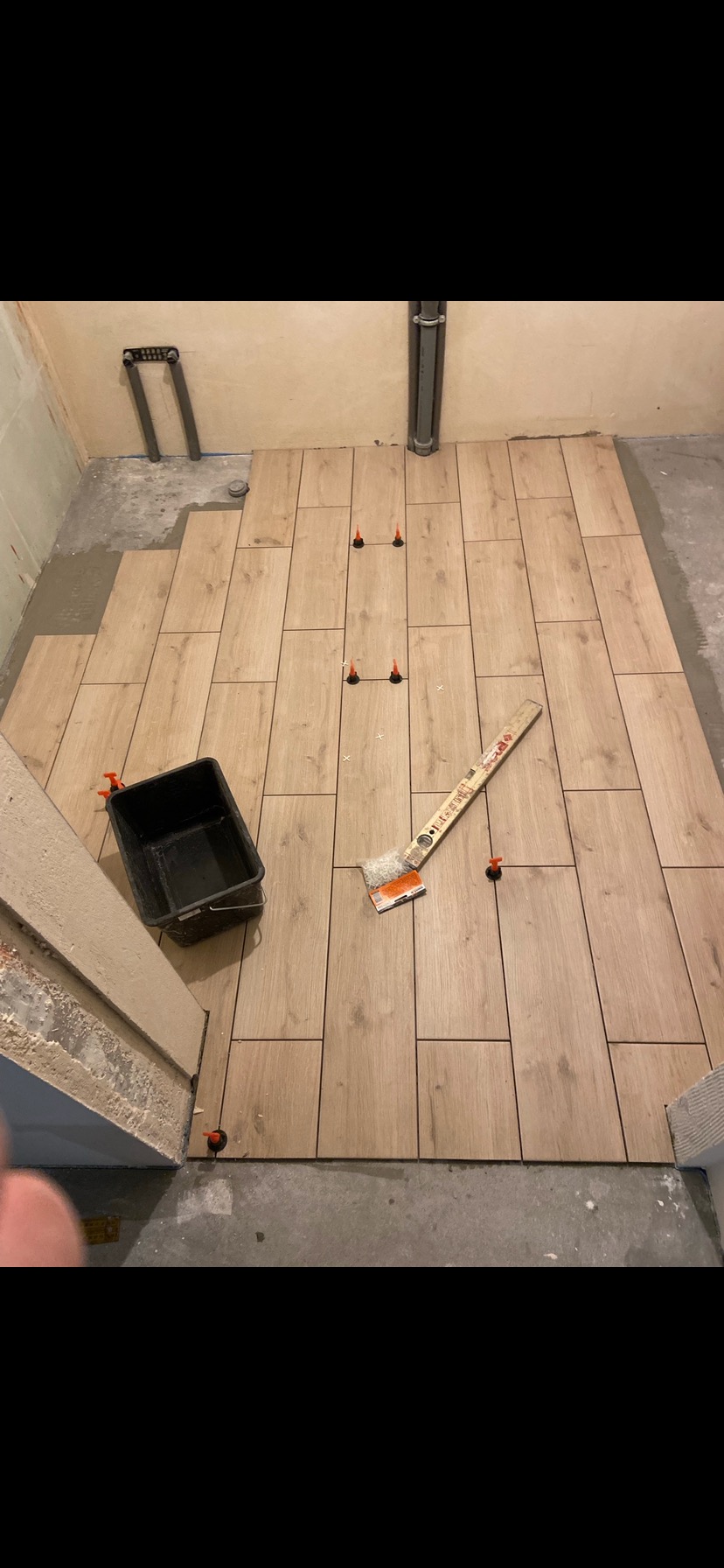 Podłoga w trakcie wykańczania: ułożone płytki imitujące drewno, poziomica, wiadro i system poziomowania płytek z pomarańczowymi klinami.