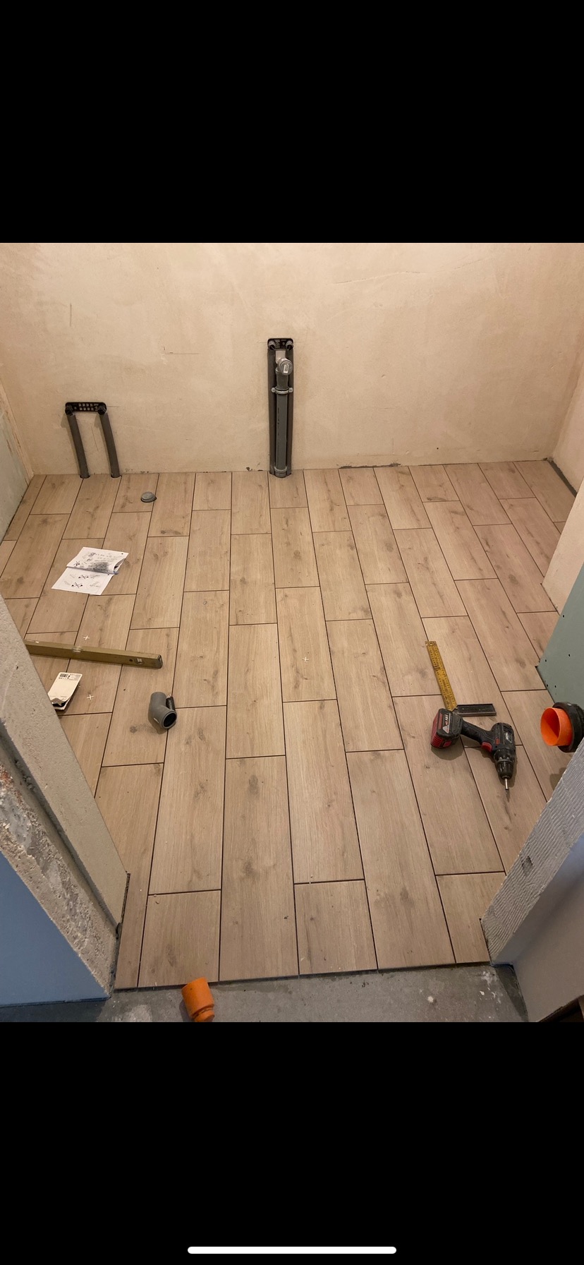 Podłoga w trakcie wykańczania z jasnych, drewnopodobnych płytek, widoczne narzędzia: poziomica, miarka, wiertarka akumulatorowa, rury kanalizacyjne i instrukcja montażu.