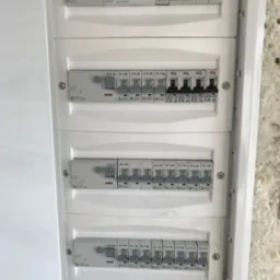 Otwarta, biała rozdzielnica elektryczna z widocznymi bezpiecznikami i wyłącznikami różnych typów, w tym zasilania głównego z sygnalizacją LED, zamontowana na jasnej, chropowatej ścianie.