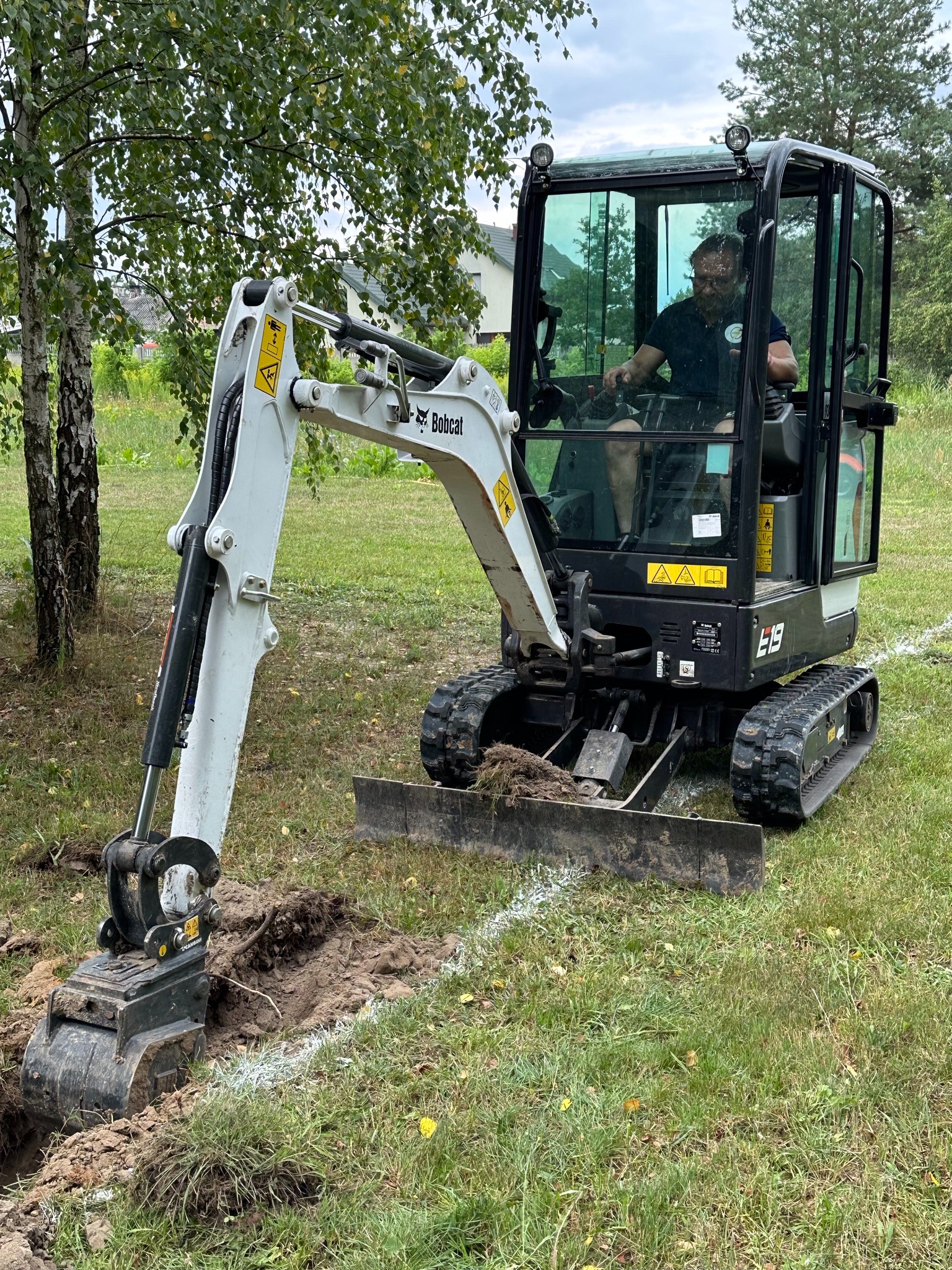 Operator w mini-koparce Bobcat E19 wykonuje wykop w ziemi, prawdopodobnie pod przyłącze, widoczne białe linie wyznaczające trasę, tło z drzewami i budynkiem.