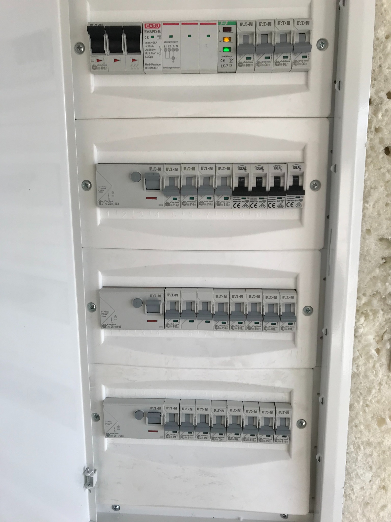 Otwarta, biała rozdzielnica elektryczna z widocznymi bezpiecznikami i wyłącznikami różnych typów, w tym zasilania głównego z sygnalizacją LED, zamontowana na jasnej, chropowatej ścianie.