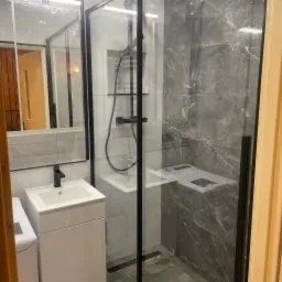 Nowoczesna kabina prysznicowa z czarnymi profilami, szare płytki imitujące marmur na ścianach, biała umywalka z szafką, lustro i widoczna pralka w tle.