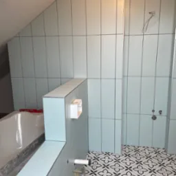 Świeżo ułożone, pionowe, jasnoniebieskie płytki ścienne z ciemnymi fugami w łazience na poddaszu. Widoczna wanna z poziomnicą, fragment ściany bez płytek, instalacja elektryczna oraz wzorzysta...