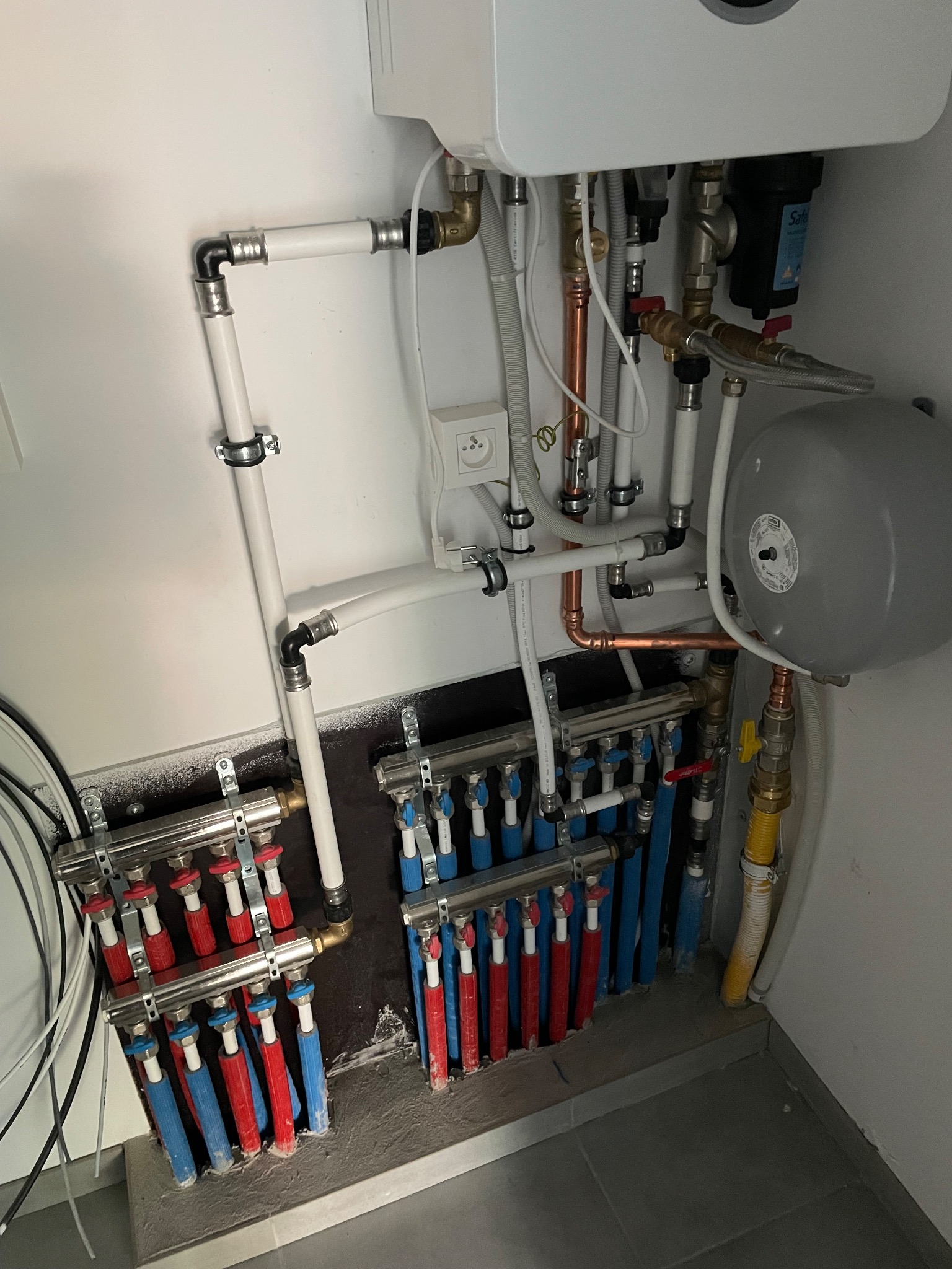 Rozdzielacz ogrzewania podłogowego z licznymi rurami, zaworami i manometrem zamontowany przy ścianie w kotłowni w Kiszkowie.