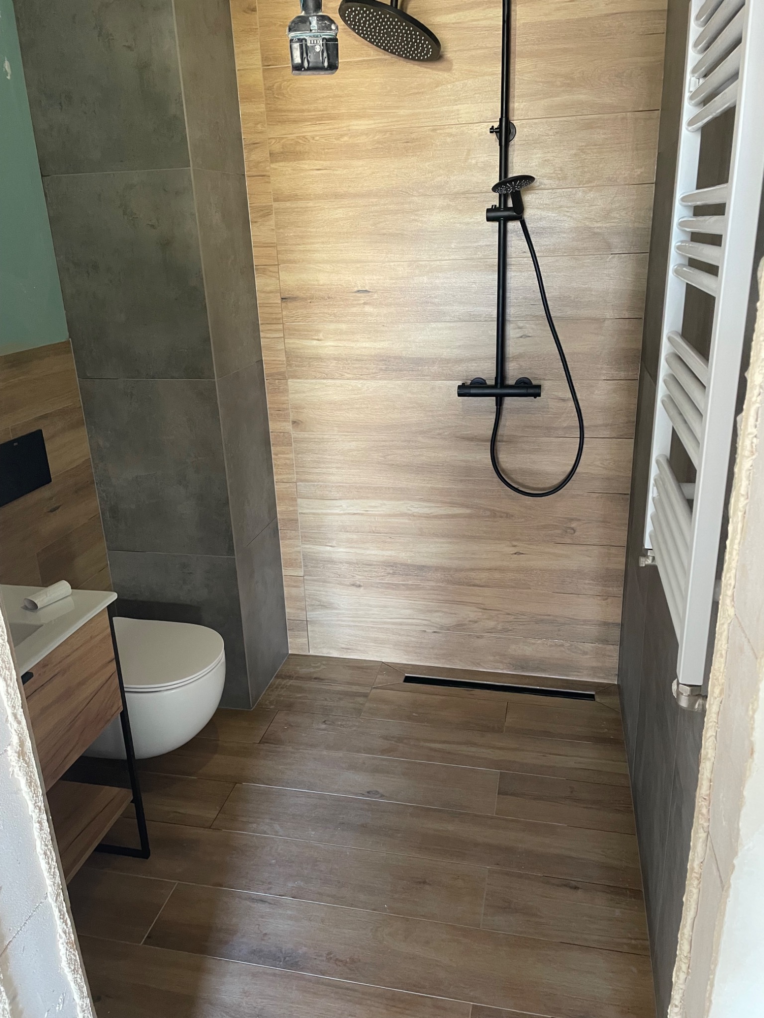 Nowoczesna łazienka z drewnopodobnymi płytkami na ścianach i podłodze, czarny zestaw prysznicowy i grzejnik drabinkowy, minimalistyczny odpływ liniowy.