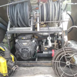 Wnętrze furgonetki z zamontowanym urządzeniem do czyszczenia kanalizacji: dwie rolki zwiniętych węży, silnik spalinowy i sprężyna kanalizacyjna na podłodze.