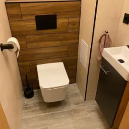 Nowoczesna toaleta podwieszana z drewnopodobną zabudową stelaża i czarnym przyciskiem spłukującym, umywalka nablatowa z szafką w ciemnym kolorze, płytki podłogowe imitujące drewno.