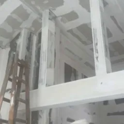 Sufit i ściany w trakcie szpachlowania płyt kartonowo-gipsowych, widoczne drewniane podpory i drabina, zabezpieczone meble folią malarską.