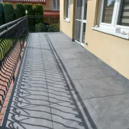 Wykończony balkon z szarymi płytkami na podłodze, czarna metalowa balustrada rzucająca wzorzysty cień na posadzkę, widok na ogród z zielonym trawnikiem i tujami.