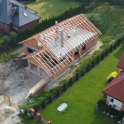 Budowa domu jednorodzinnego w stanie surowym otwartym, widok z góry, z widoczną konstrukcją dachu i ścianami z cegły.