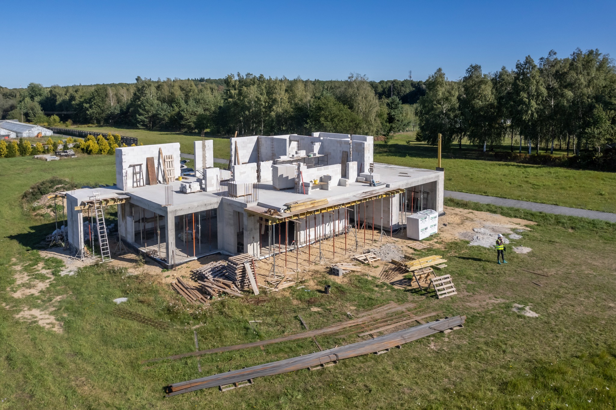 Surowy stan domu z betonu komórkowego w trakcie budowy, widok z góry. Widoczny pracownik w kasku i elementy konstrukcyjne, szalunki, zbrojenia. W tle zieleń.