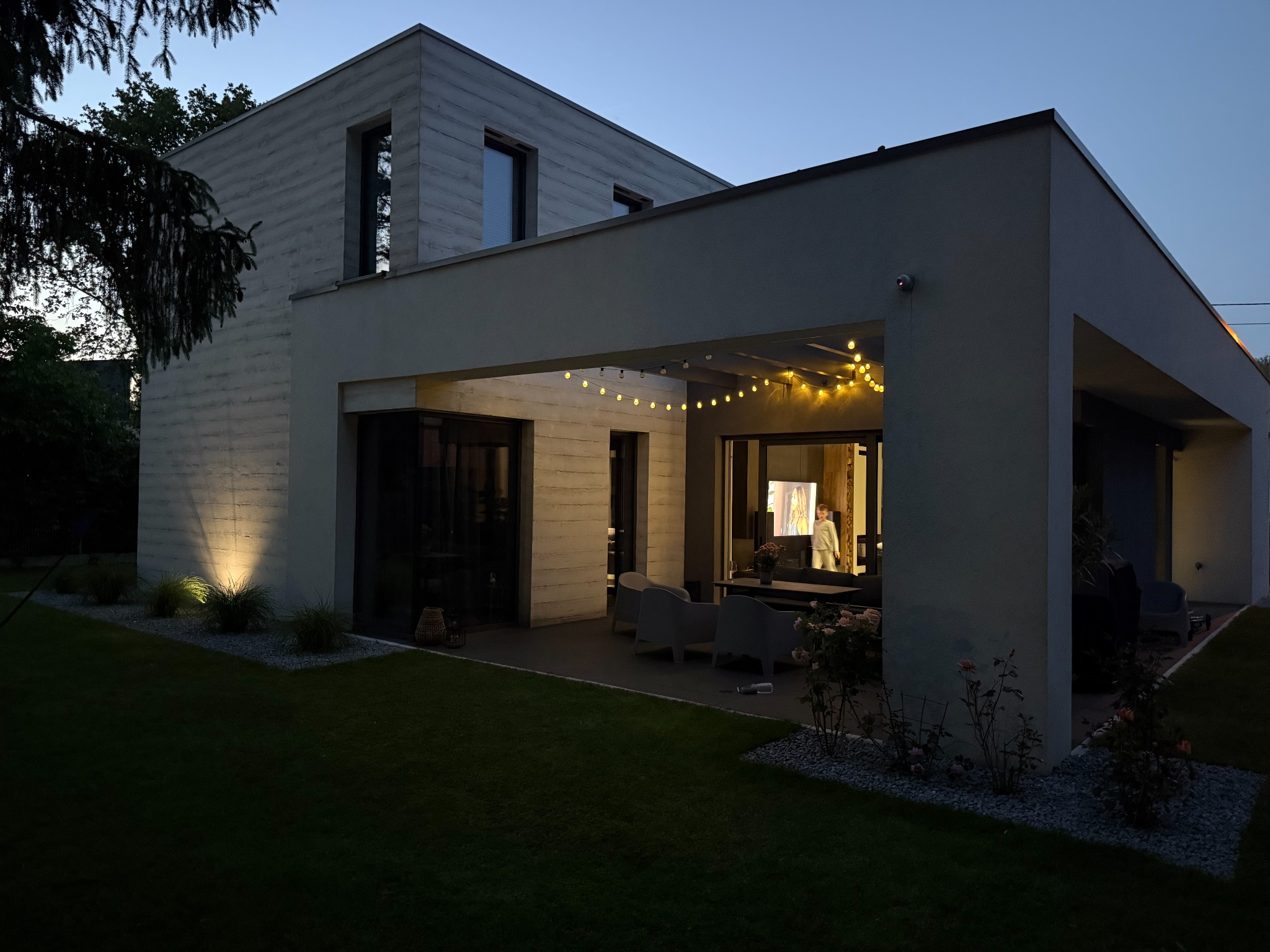Nowoczesny dom z oświetlonym tarasem i ogrodem o zmierzchu. Minimalistyczna architektura, ciepłe światło girland i lamp podkreśla detale elewacji.