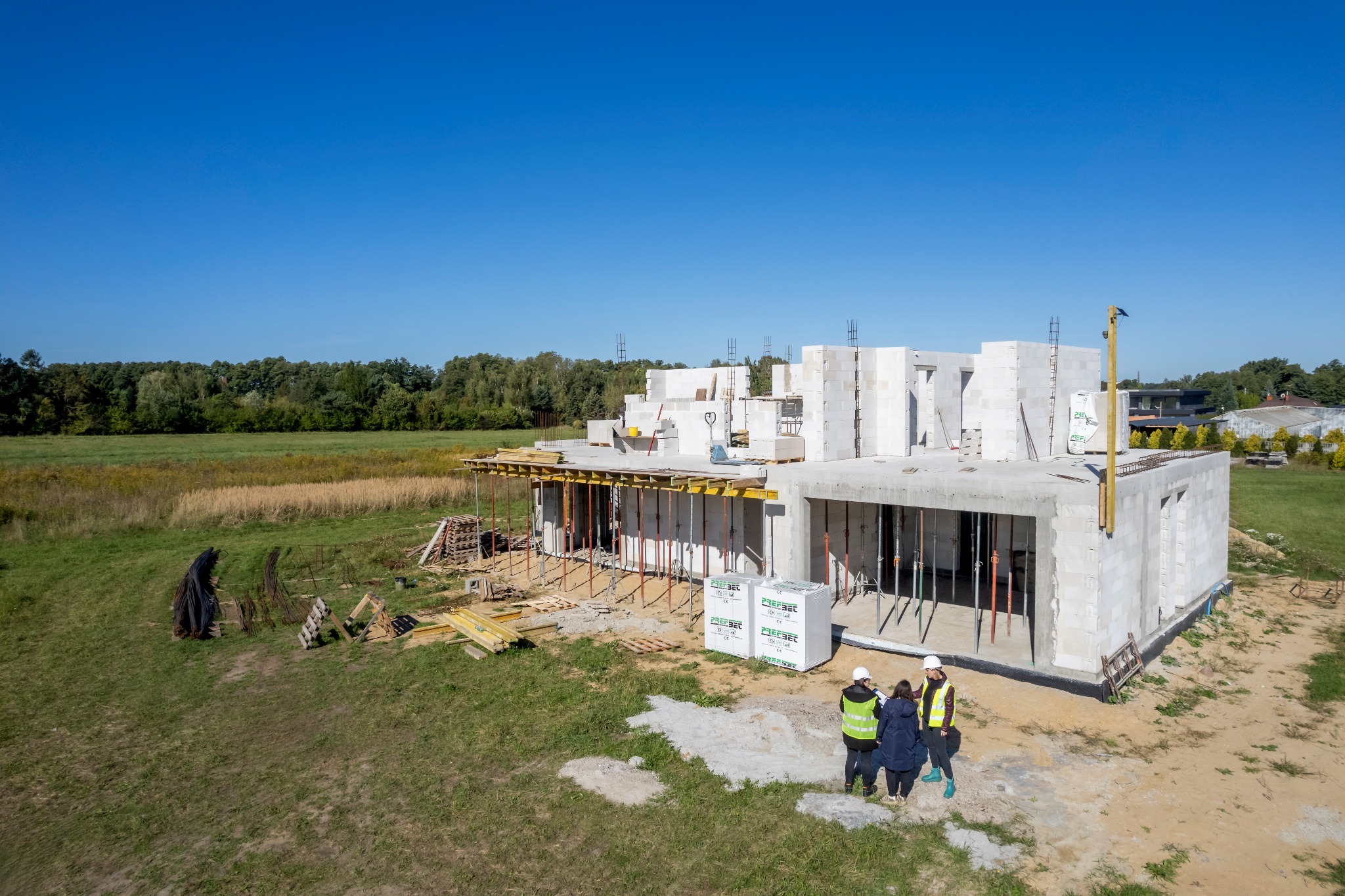 Budowa domu z betonu komórkowego w trakcie realizacji. Widoczne ściany, strop, szalunki i osoby w kaskach omawiające projekt na tle zieleni i błękitnego nieba.