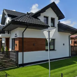 Nowoczesny dom jednorodzinny z białą elewacją, elementami drewnianymi i czarnym dachem, widoczny z perspektywy ulicy w słoneczny dzień, z zadbanym trawnikiem i znakiem drogowym na pierwszym planie.