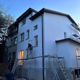 POL - DEK Sławomir Polesiak - Dwupiętrowy budynek jednorodzinny z nową, jasnoszarą elewacją, pracownik na rusztowaniu w trakcie prac wykończeniowych, widoczne narzędzia i materiały budowlane, wieczorny krajobraz.