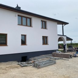 POL - DEK Sławomir Polesiak - Dwukondygnacyjny dom z jasną elewacją i brązowymi oknami, widok z boku, z widocznymi rusztowaniami i materiałami budowlanymi na placu budowy.