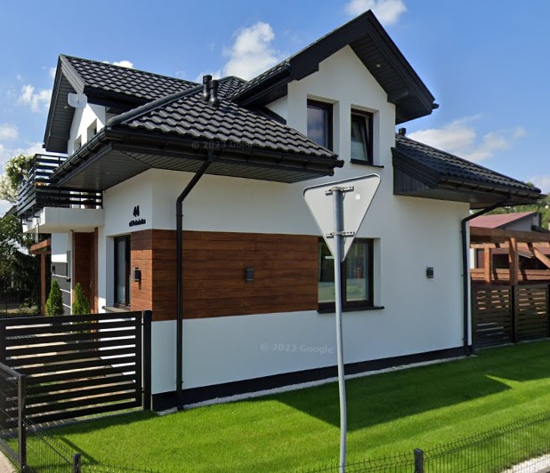 Nowoczesny dom jednorodzinny z białą elewacją, elementami drewnianymi i czarnym dachem, widoczny z perspektywy ulicy w słoneczny dzień, z zadbanym trawnikiem i znakiem drogowym na pierwszym planie.