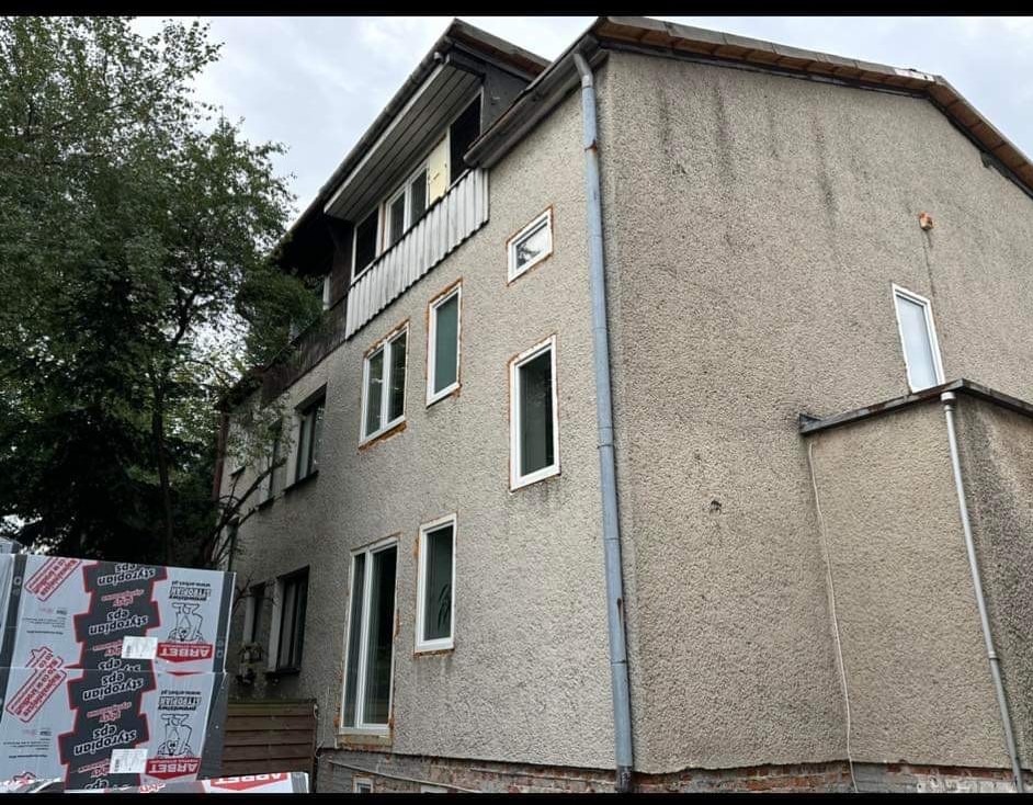 Dwupiętrowy budynek z szarą elewacją przed rozpoczęciem prac. Widoczne paczki styropianu na pierwszym planie, przygotowane do ocieplenia.