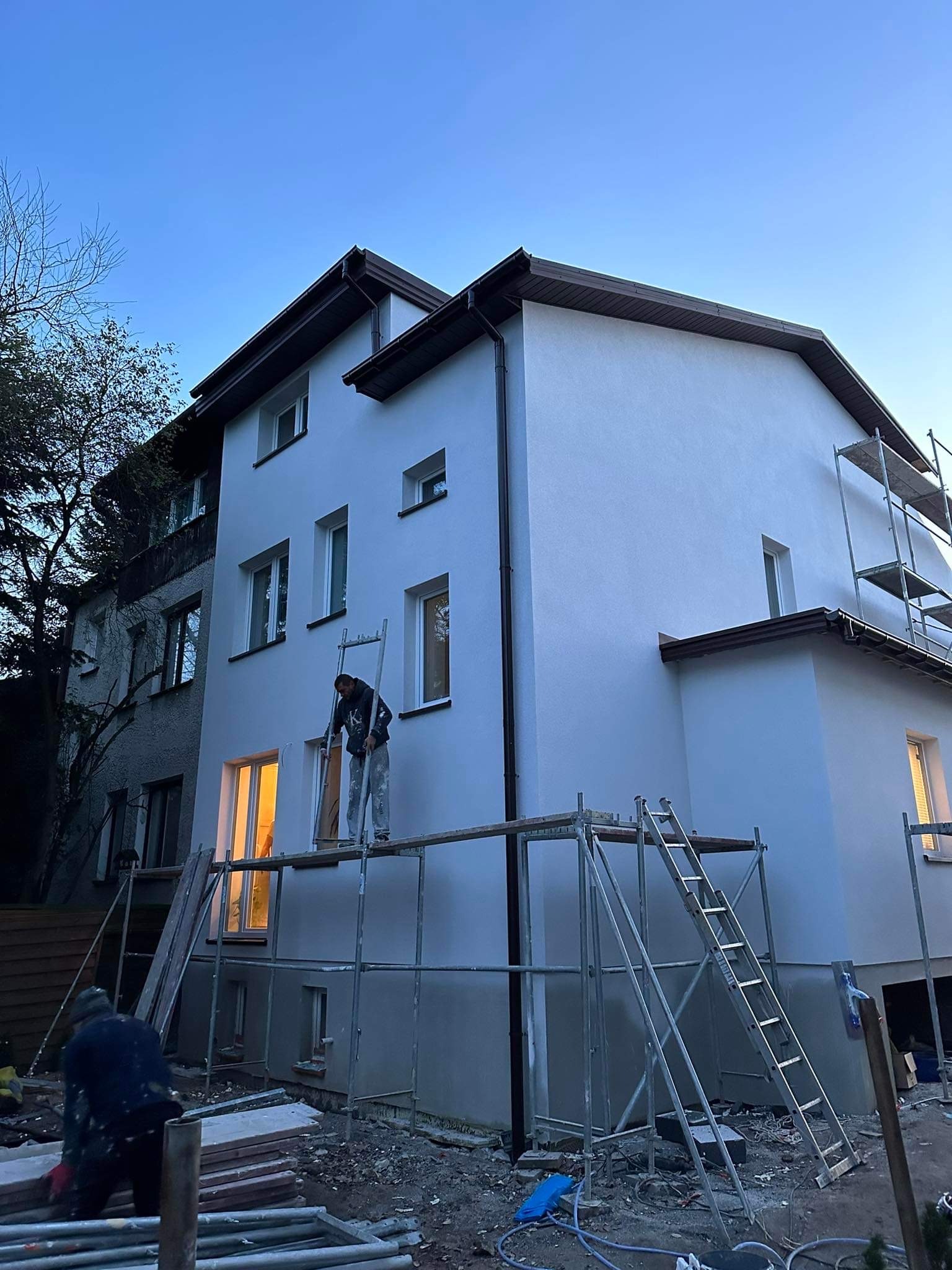 Dwupiętrowy budynek jednorodzinny z nową, jasnoszarą elewacją, pracownik na rusztowaniu w trakcie prac wykończeniowych, widoczne narzędzia i materiały budowlane, wieczorny krajobraz.