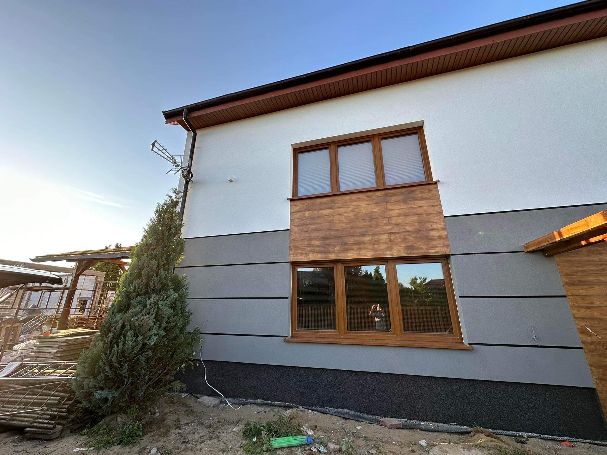 Dwukondygnacyjny dom z szarą elewacją podzieloną poziomymi pasami, drewnianymi panelami pod oknami i ciemną, ziarnistą podstawą, widok z dołu, antena na ścianie, iglak przy ścianie.