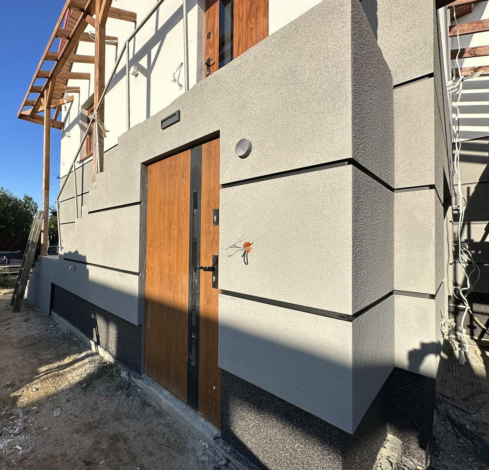 Szara elewacja domu jednorodzinnego w Białobrzegach z drewnianymi drzwiami wejściowymi, widoczna struktura tynku i fragment izolacji fundamentów, w tle rusztowanie.