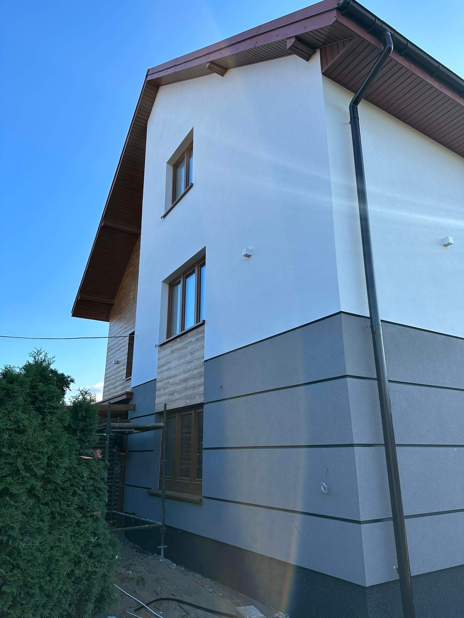 Dwukondygnacyjny dom z elewacją w odcieniach bieli i szarości, z widocznymi rusztowaniami przy ścianie bocznej, brązowe okna i dachówka, na tle błękitnego nieba.