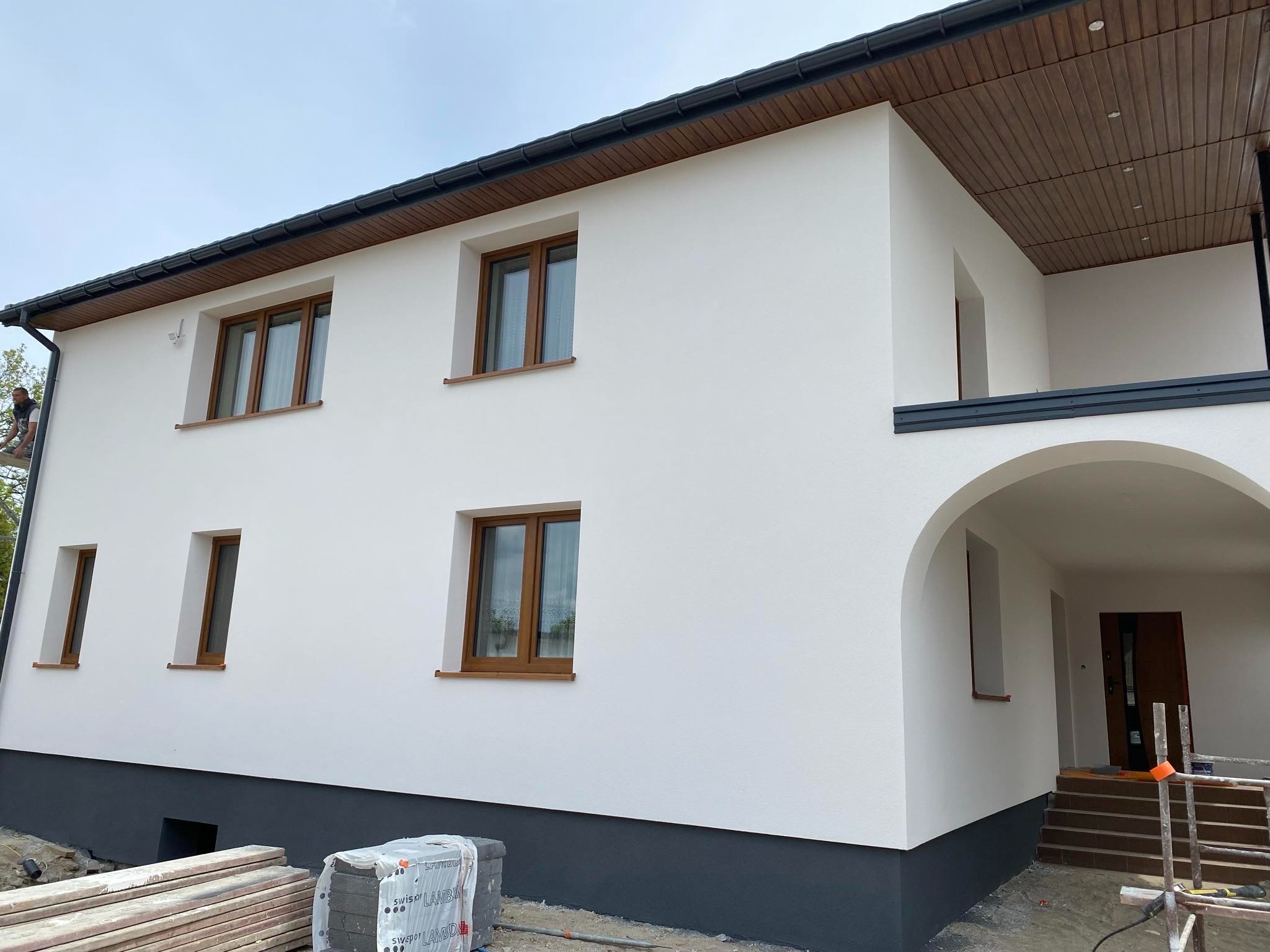 Dwukondygnacyjny dom z białą elewacją i brązowymi oknami, widoczne przygotowane materiały budowlane u podstawy ściany, oraz zadaszone wejście z łukiem.