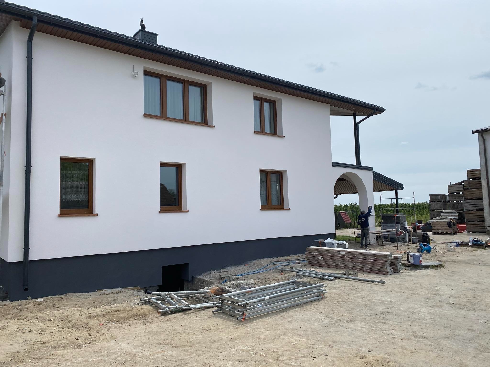 Dwukondygnacyjny dom z jasną elewacją i brązowymi oknami, widok z boku, z widocznymi rusztowaniami i materiałami budowlanymi na placu budowy.