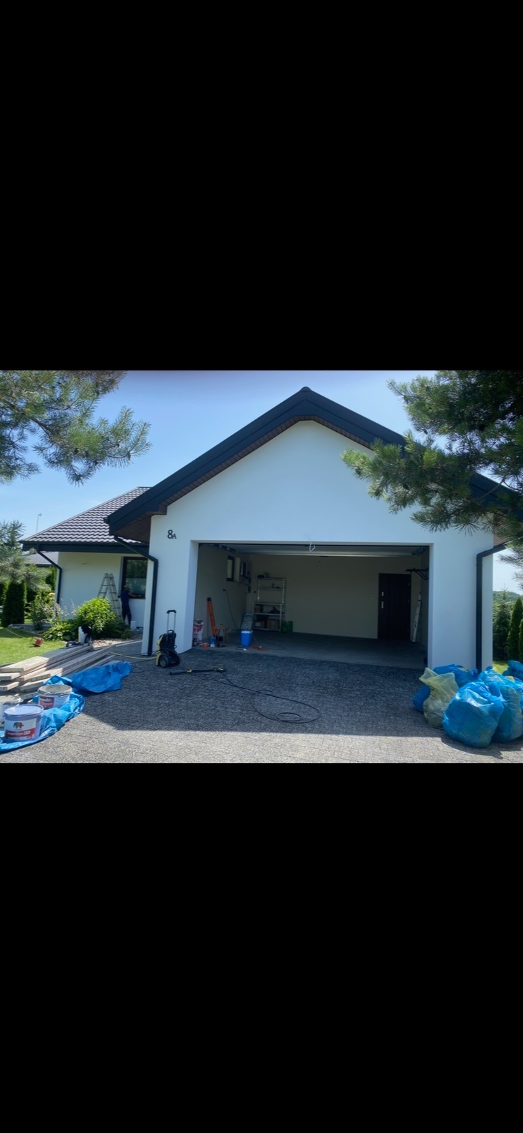 Dom jednorodzinny z otwartym garażem podczas prac wykończeniowych elewacji, widoczne farby, narzędzia i worki na odpady budowlane na podjeździe.