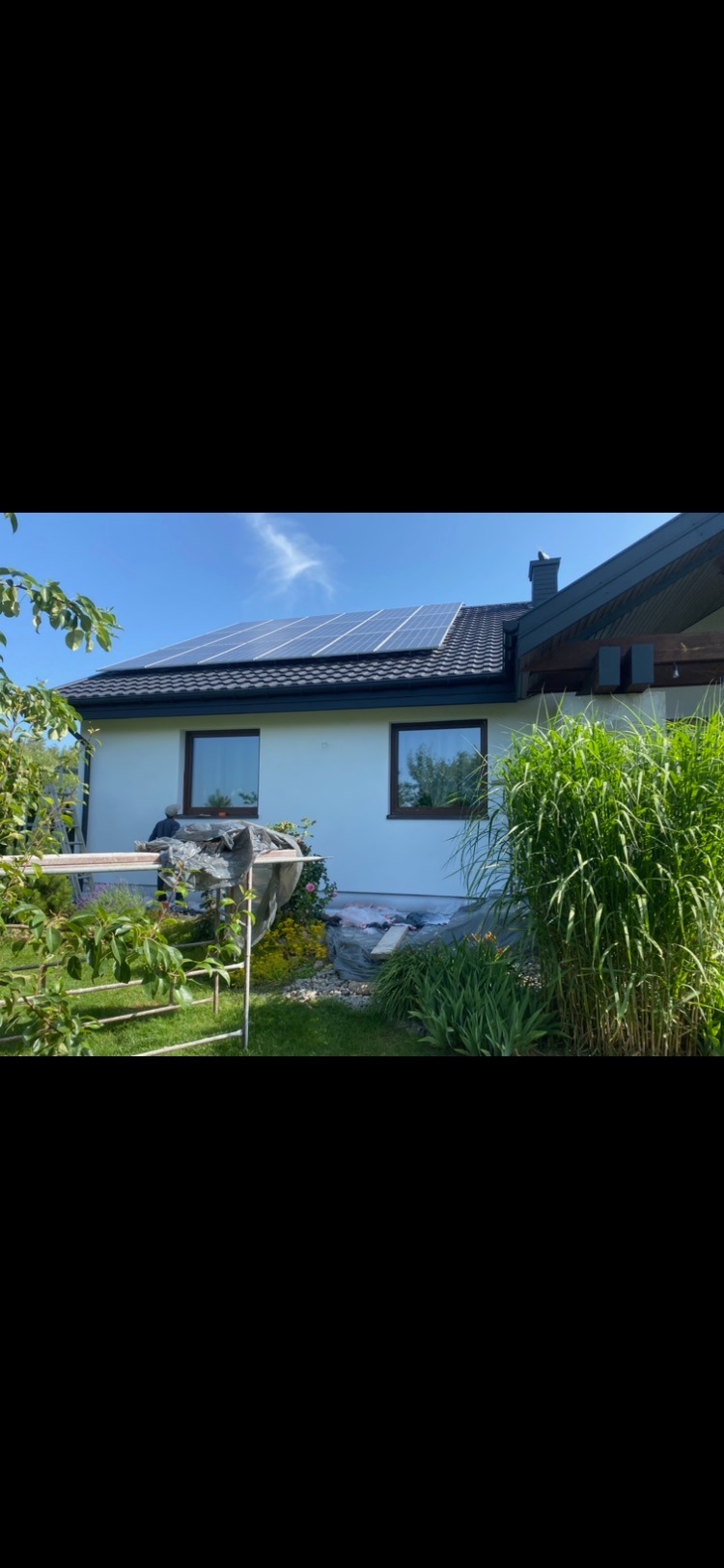 Biały dom jednorodzinny z panelami słonecznymi na dachu, widoczne okna z brązowymi ramami i fragment ogrodu z zielenią.