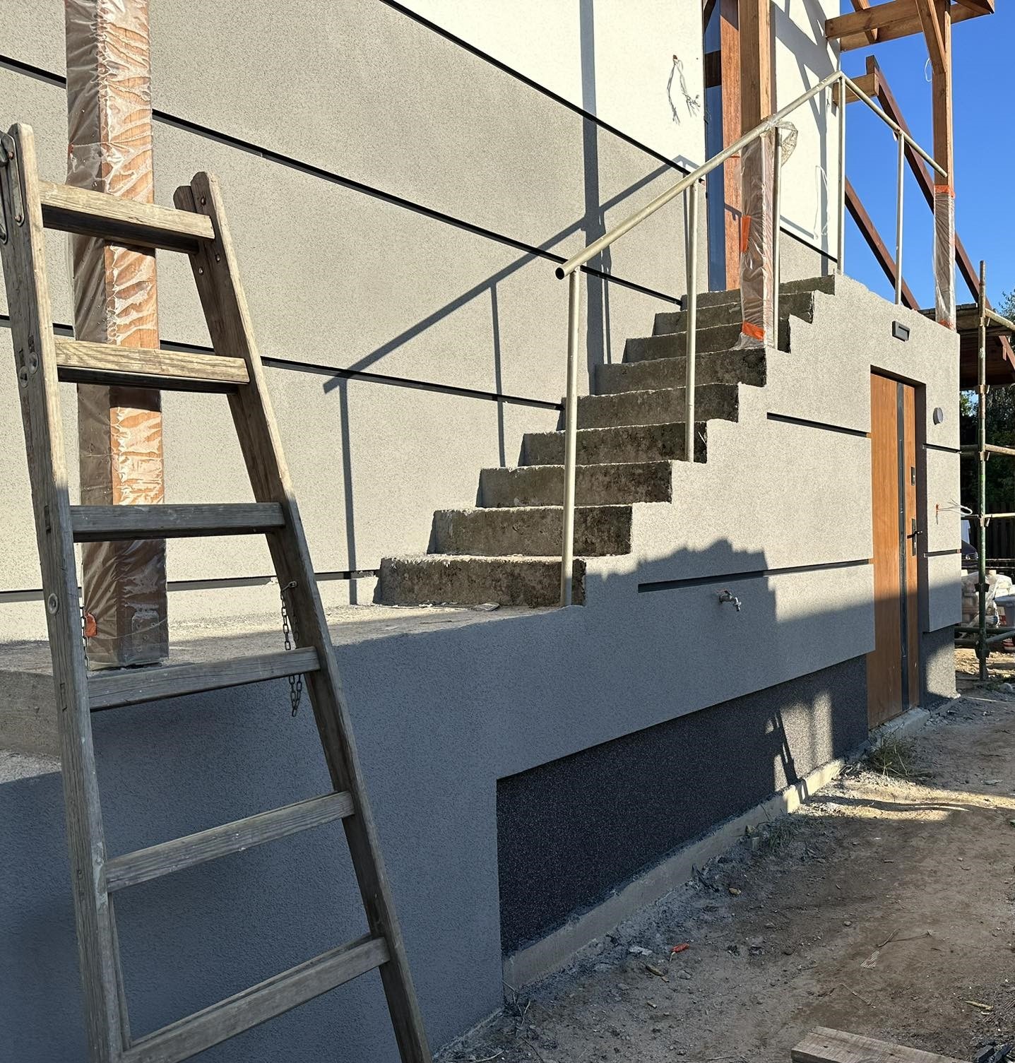 Szara elewacja budynku z widocznymi schodami zewnętrznymi z metalową barierką, drewnianą drabiną opartą o ścianę oraz fragmentami rusztowania i niezabezpieczonym słupem, ukazująca detale tynku...