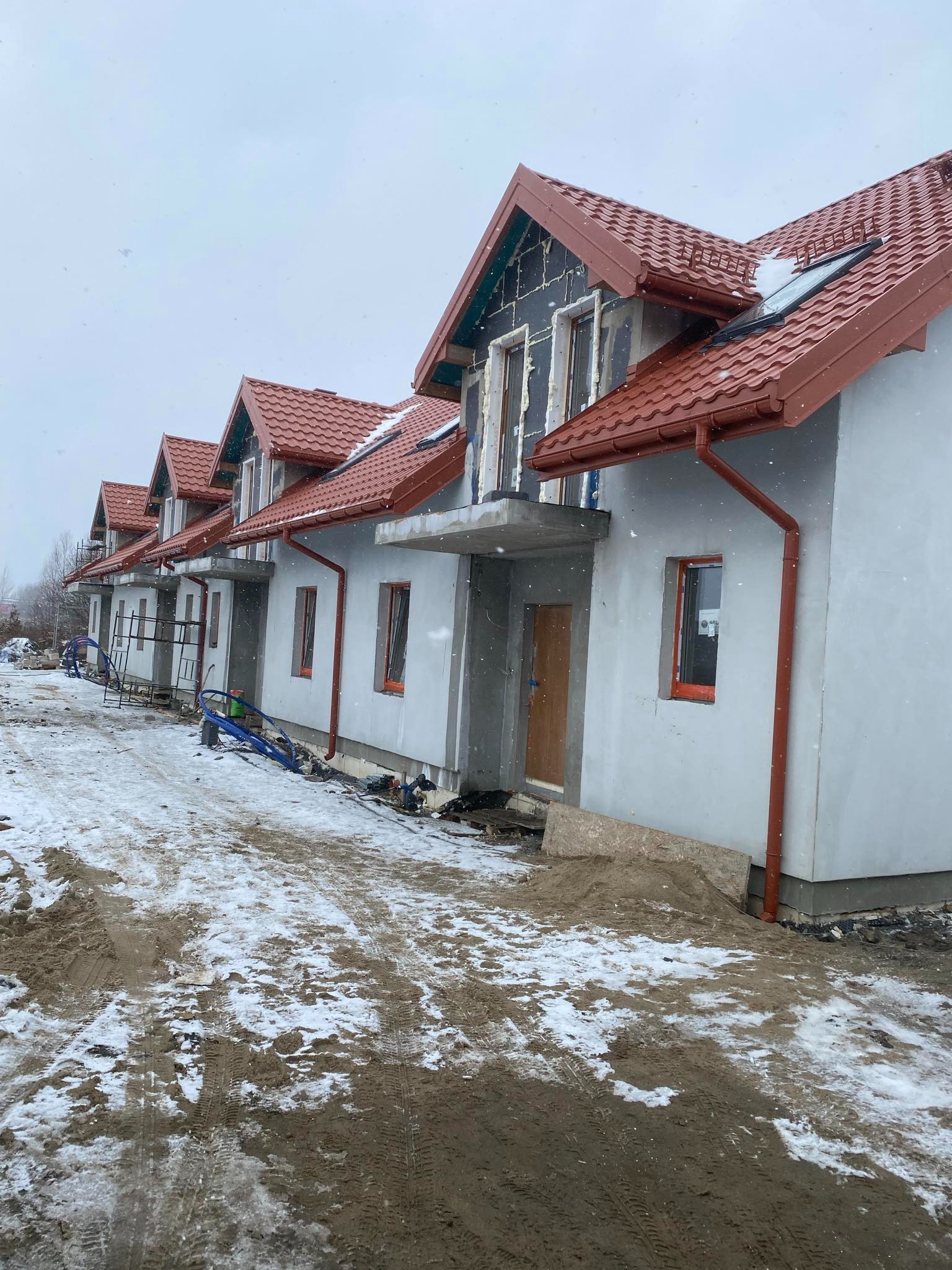 Szereg nowo budowanych domów jednorodzinnych w stanie surowym zamkniętym, z czerwoną dachówką, orynnowaniem i częściowo otynkowanymi elewacjami, w zimowej scenerii z padającym śniegiem.