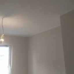 Świeżo pomalowane, beżowe ściany i sufit w pustym pokoju, widoczne przewody oświetleniowe, listwa przysufitowa i okno z workiem foliowym.