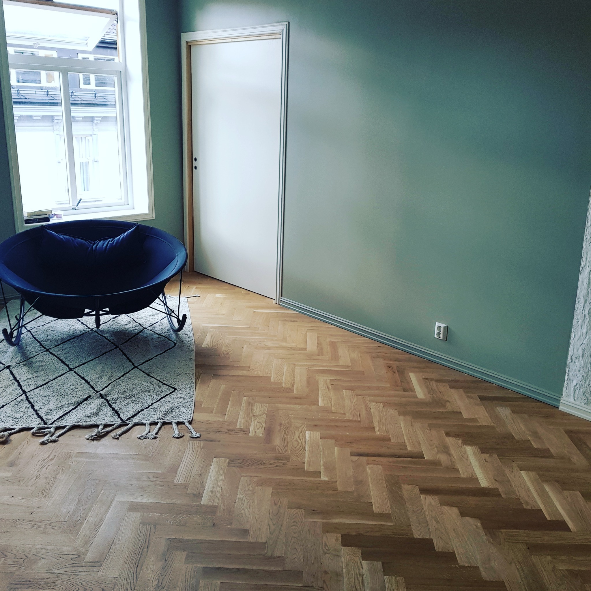 Jasny parkiet ułożony w jodełkę francuską, świeżo pomalowane ściany na kolor szałwiowej zieleni, białe drzwi i okno z widokiem na budynek naprzeciwko, fotel bujany z poduszką i dywanik z frędzlami.