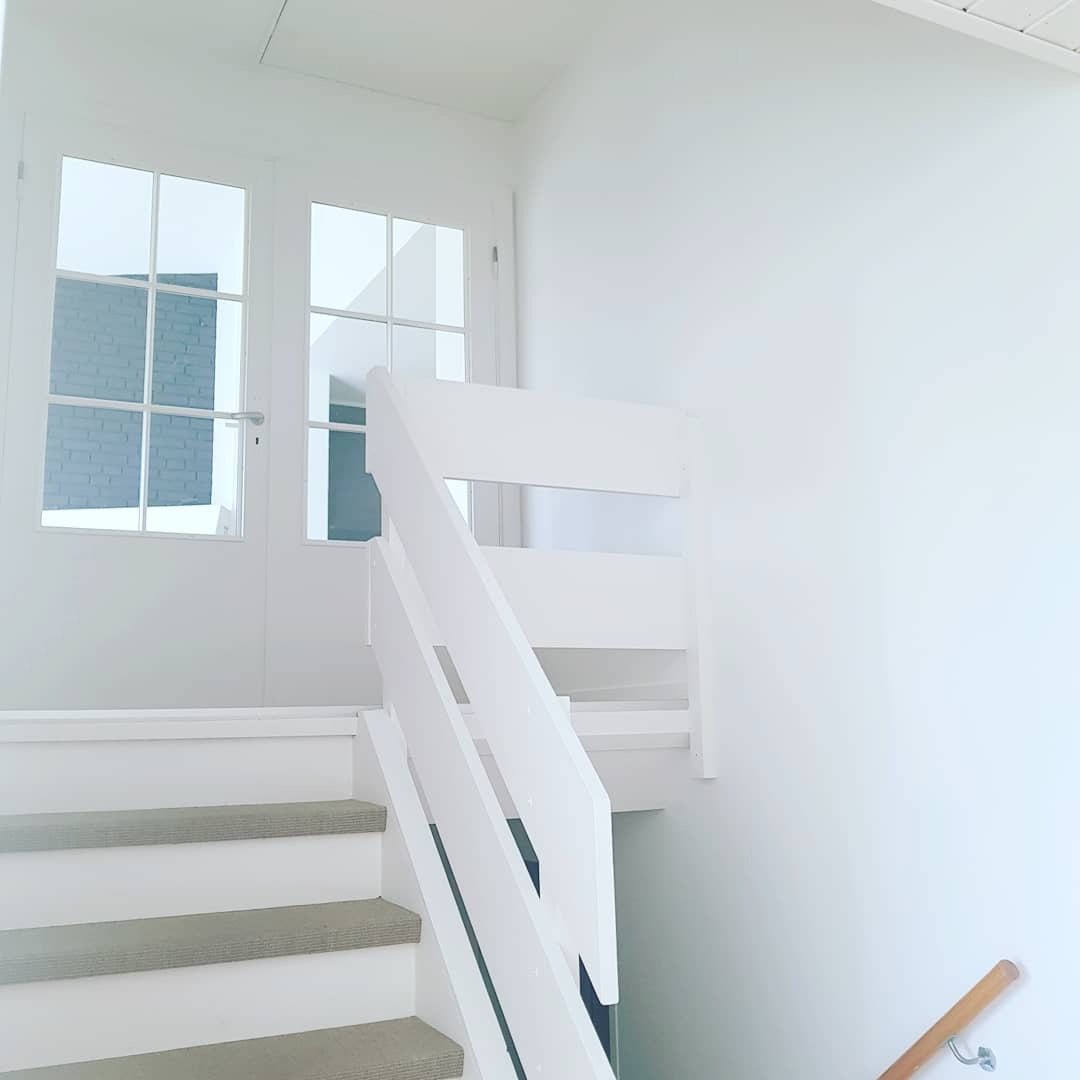 Wnętrze domu po odświeżeniu: białe ściany, schody z jasną wykładziną i biała balustrada, przeszklone drzwi wejściowe z widokiem na ceglany mur.